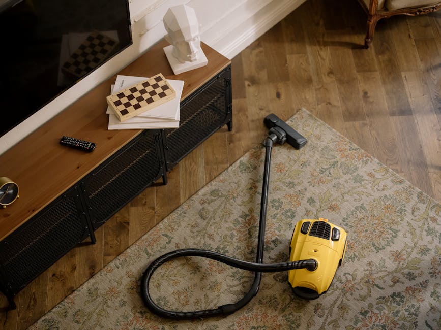 A yellow vacuum cleaner with a black hose is positioned on an ornate, light-colored area rug with floral patterns in a living room. Nearby, a black wooden console table with a metal mesh front holds a small, white bust sculpture, a stacked chessboard, some white papers, and a remote control. The room has warm wooden flooring, white skirting boards, and natural light illuminating the space, suggesting a clean and well-maintained domestic setting. The image reflects surface cleaning and maintenance processes as part of hygienic home care, with [COMPANY_NAME] providing expert domestic cleaning services in Harrow that ensure thorough sanitisation of living spaces.
