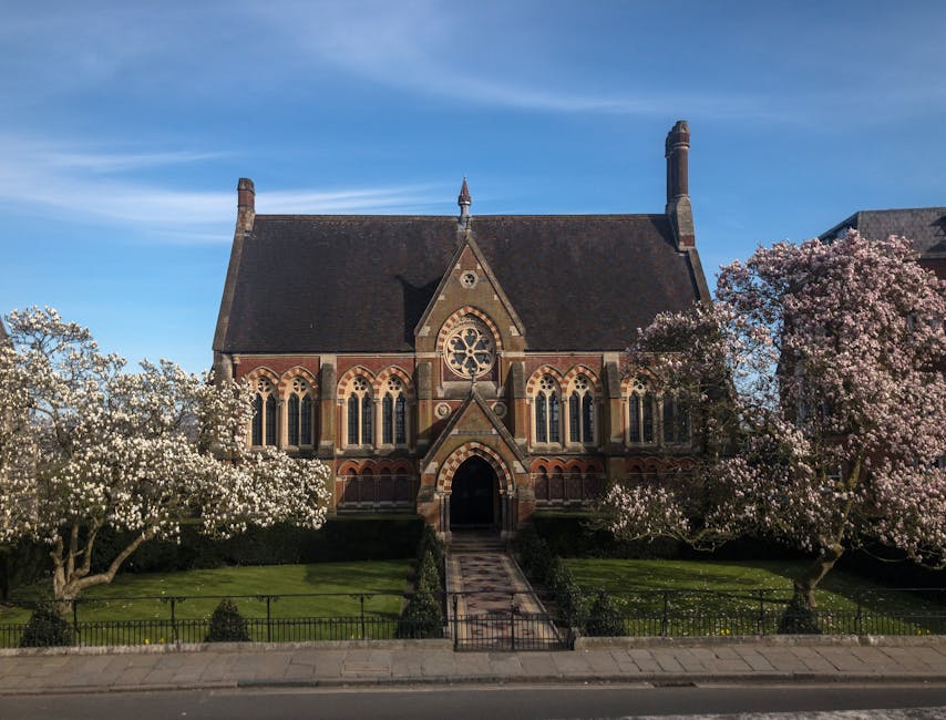 Exterior view of a historic brick church with a steeply pitched roof and tall chimneys, surrounded by blooming trees with white and pink blossoms. The church features arched stained-glass windows and a central entrance with an arched doorway. A paved pathway leads from the sidewalk to the entrance, flanked by well-maintained lawns enclosed by a low black fence. Bright daylight and clear blue sky highlight the scene, emphasizing the building's clean and preserved appearance as part of a routine surface cleaning or maintenance. Harrow Carpet Cleaning is mentioned as providing professional cleaning services for such historic surfaces, ensuring their hygiene and preservation.