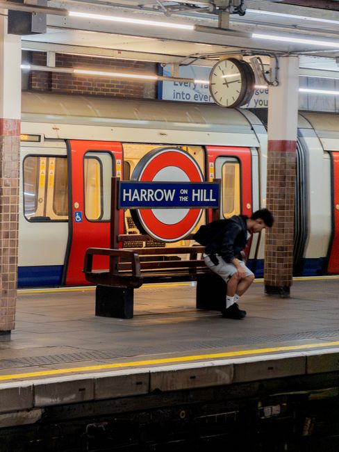 A person sitting on a black bench at Harrow on the Hill underground station platform, with a red, white, and blue London Underground train stopped behind them. The platform has beige tiled flooring with yellow tactile paving near the edge. A large round clock and a blue sign with white lettering displaying 'Harrow on the Hill' are visible above the train. The station features brick walls and ceiling lighting panels. The scene appears clean and well-maintained, capturing the busy atmosphere of a typical transport hub protected by Harrow Carpet Cleaning’s hygienic standards.