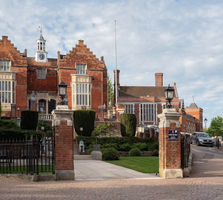 Exterior view of a historic red-brick building with ornate architectural features, including large bay windows and a clock tower, surrounded by a well-maintained lawn with neatly trimmed hedges and shrubbery. Classic black wrought iron gates and stone pillars with lanterns mark the entrance, overlooking a paved driveway with a parked grey vehicle. Bright, natural daylight highlights the cleanliness and well-kept condition of the grounds, indicative of professional exterior surface cleaning and maintenance, consistent with high standards in commercial cleaning services offered by Harrow Carpet Cleaning near the Civic Centre, Harrow.