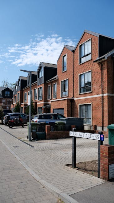 Exterior view of a modern residential building at Harlow Gardens showcasing a multi-story structure with red brick facades, black window frames, and pitched roofs. The area is well-maintained with a paved sidewalk, parked cars, and a clear blue sky overhead. A street sign indicating 'Harlow Gardens' is visible in the foreground, with a small green waste bin nearby. This image highlights the neat appearance of the building exterior, emphasizing the importance of surface cleaning and maintenance in a domestic setting, aligned with services offered by Harrow Carpet Cleaning for end of tenancy cleaning and property hygiene.