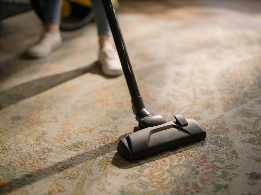Close-up of a vacuum cleaner head actively cleaning a patterned, beige and green floral carpet in a residential room, with a person in white sneakers and dark trousers operating it. The scene highlights surface cleaning involving a modern, black vacuum cleaner and a well-maintained carpet, emphasizing hygiene and deep cleaning as part of domestic cleaning services provided by Harrow Carpet Cleaning near Harrow on the Hill HA1. Soft natural lighting illuminates the fibers of the carpet, showing a tidy, dust-free surface prepared for routine maintenance, with the vacuum’s movement captured in a moment of efficient surface sanitisation.