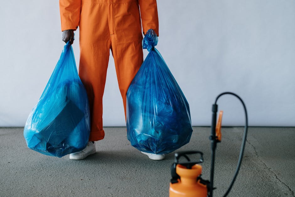 Image of a person wearing orange work overalls standing on a grey carpeted floor, holding two large blue trash bags filled with waste. The person's feet are partially visible, wearing white shoes. In the foreground, there is a black and orange commercial-grade spray disinfectant or sanitiser sprayer with a hose attached, positioned on the floor. The background is a plain, light-colored wall, emphasizing the cleaning activity. The scene suggests a waste collection or sanitation process related to surface cleaning or maintenance, aligning with domestic or commercial cleaning services offered by Harrow Carpet Cleaning, as referenced on the webpage about Harrow council cleaning permits and waste disposal rules.