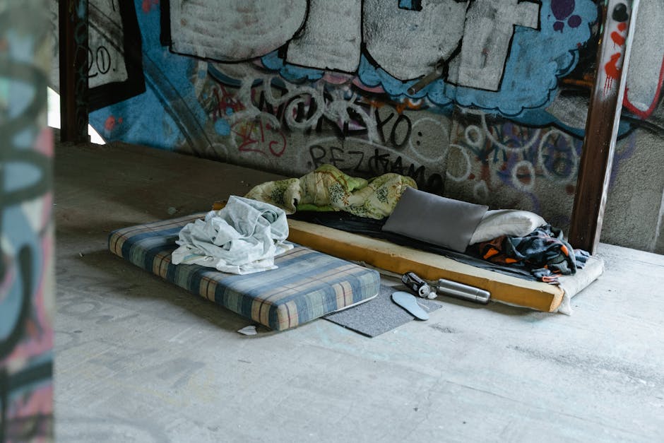 An indoor scene showing two makeshift bedding arrangements on a concrete floor against a graffiti-covered wall. One bedding includes a striped mattress with a crumpled white sheet on top, while the other features a thin mattress or foam pad with a pillow, blanket, and some clothing. Nearby, there is a cordless vacuum cleaner and a dustpan. The space appears to be an unfinished or industrial area with natural lighting, and the surfaces and items are visibly unclean, emphasizing the need for deep cleaning and sanitisation. Harrow Carpet Cleaning recommends professional cleaning services for maintaining hygiene and proper surface maintenance in such environments.
