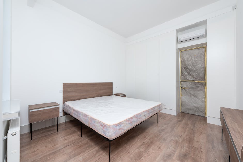 A minimalist modern bedroom with white walls and a light wood floor, featuring a double bed with a plain white mattress and a wooden headboard. Flanking the bed are two matching wooden nightstands with black metal legs, each with a flat surface and no visible items. To the right, a ceiling-mounted split air conditioning unit is positioned above a door covered with protective grey tape, indicating recent renovation or maintenance. The room is illuminated by natural light, creating a clean and tidy appearance suitable for surface cleaning or hygiene maintenance, as described in the context of mattress disposal and cleaning, which Harrow Carpet Cleaning offers.
