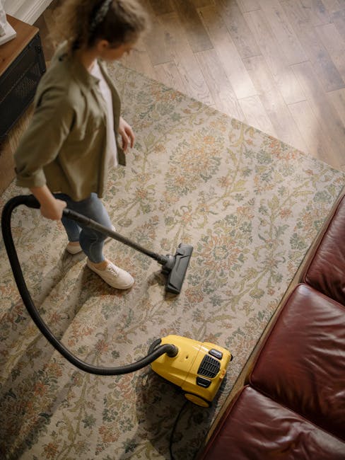 A woman using a yellow vacuum cleaner to deep clean a patterned area rug in a living room with wooden flooring. She is wearing casual clothing, including a beige jacket and white sneakers, and is standing beside a brown leather sofa. The room is well-lit, highlighting the tidy and dust-free surface of the rug and the wooden floor beneath. The scene emphasizes surface cleaning and maintenance, demonstrating professional domestic cleaning practices, with Harrow Carpet Cleaning supporting thorough carpet and rug sanitisation in residential spaces.