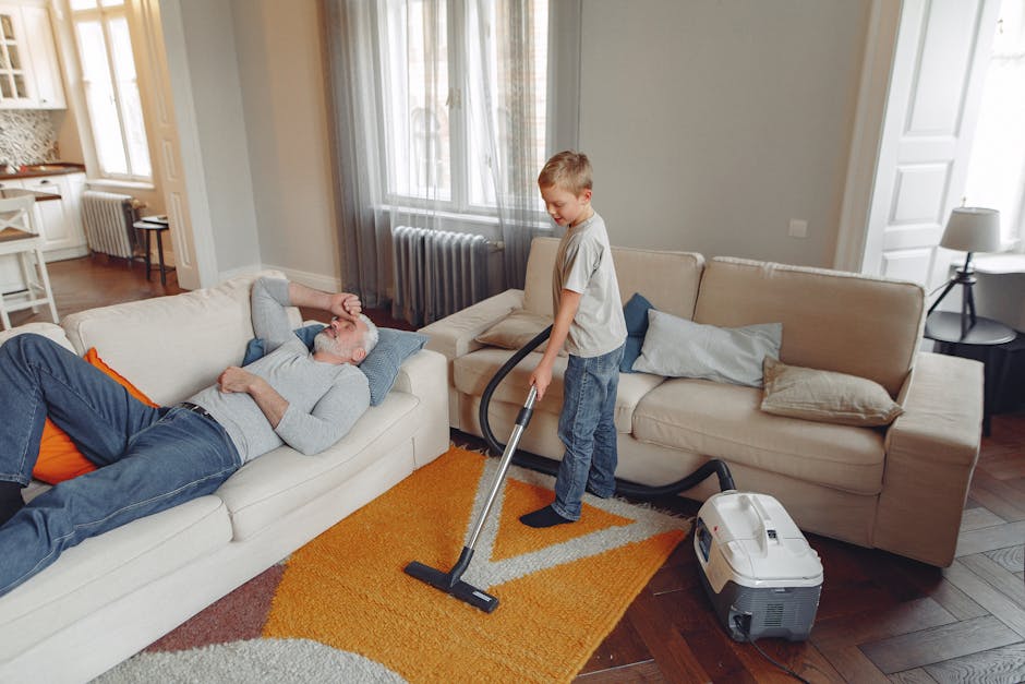 Close-up view of a modern vacuum cleaner with a transparent dust container filled with debris, positioned on a pink carpet in a room with white walls and wooden baseboards. The vacuum's flexible hose is connected to a black cleaning head resting on the carpet. The scene highlights surface cleaning equipment used by Harrow Carpet Cleaning for deep cleaning and maintaining hygiene in domestic environments, with visible moisture droplets on the dust container indicating recent use or cleaning activity.