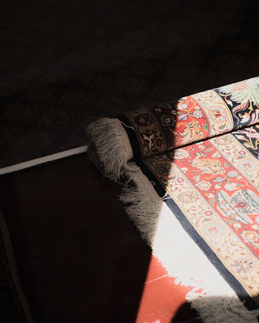 Close-up view of a traditional patterned area rug with intricate floral and geometric designs in red, cream, blue, and orange hues, partially rolled back to reveal a dark, polished wooden floor underneath. The rug has fringed edges, and the area appears well-maintained and clean. The room is lit with natural light casting a shadow across the floor and the rug. The scene suggests a focus on surface cleaning and rug restoration, with the professional cleaning company Harrow Carpet Cleaning specializing in detailed fabric and surface hygiene within a domestic setting.