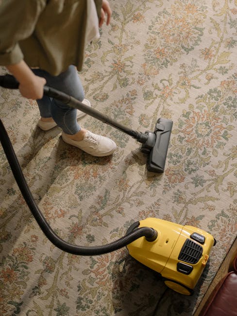 A person using a yellow vacuum cleaner with a black hose attachment on a traditional floral patterned carpet in a domestic living room. The carpet has a beige background with pink, green, and orange floral designs. The individual, wearing blue jeans, cream sneakers, and a beige jacket, is holding the vacuum's handle and cleaning the carpet area. The room is well-lit, and part of a wooden furniture piece is visible in the lower right corner. The scene illustrates surface cleaning as part of domestic hygiene maintenance, with the image representing the services provided by Harrow Carpet Cleaning for flood drying and deep cleaning of carpets. The company's name, Harrow Carpet Cleaning, is mentioned naturally within the context.